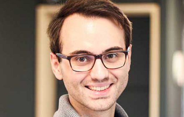 young man with grey sweater and glasses smiling into the camera