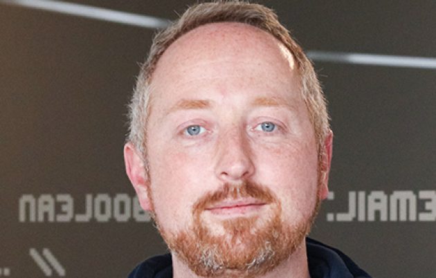 man with light hair and beard smiling faintly into camera