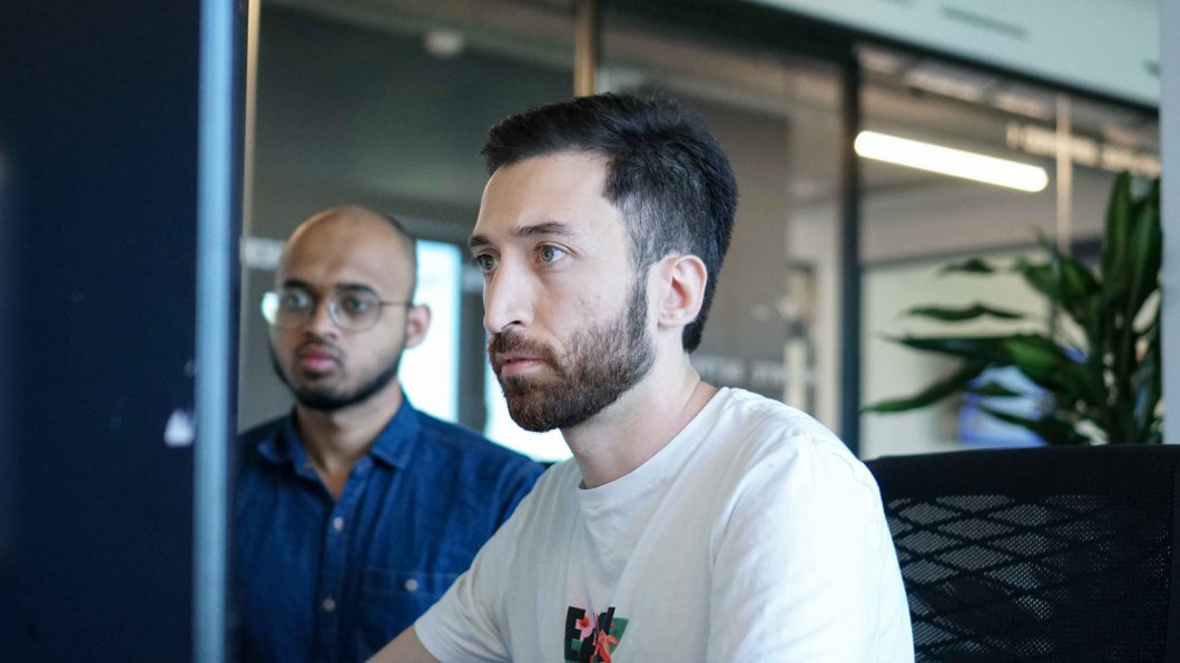 One young tech employee sitting down in front of a computer screen next to another colleague looking serious
