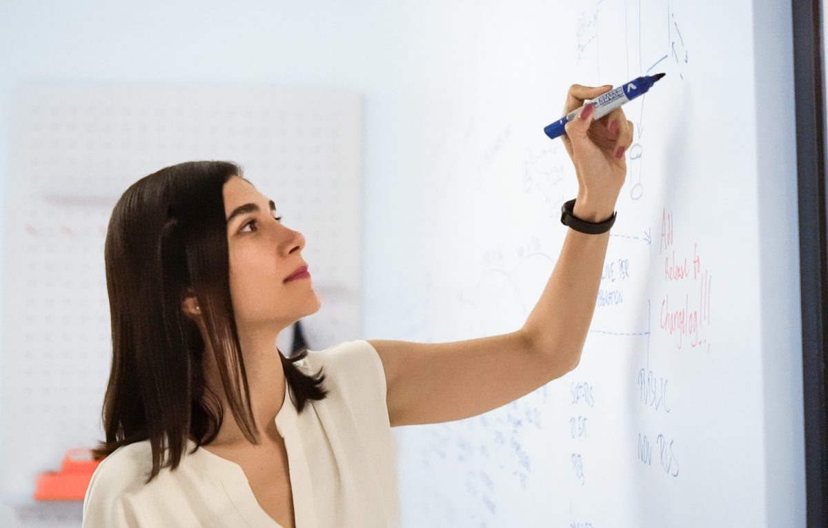 young woman with dark hair uses a marker to write on the whiteboard