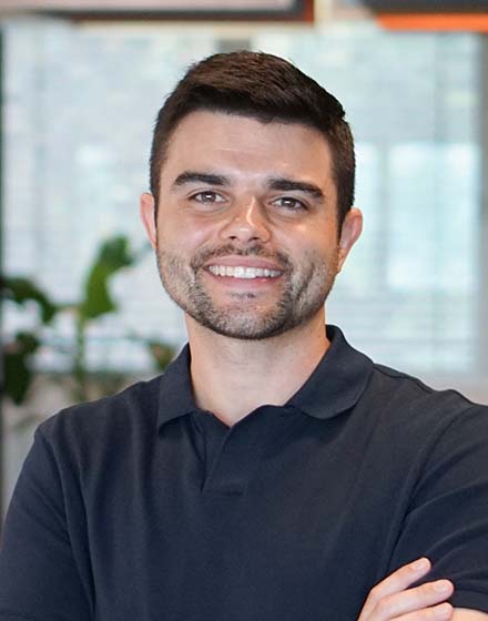 young dark-haired man with office behind him smiling at camera