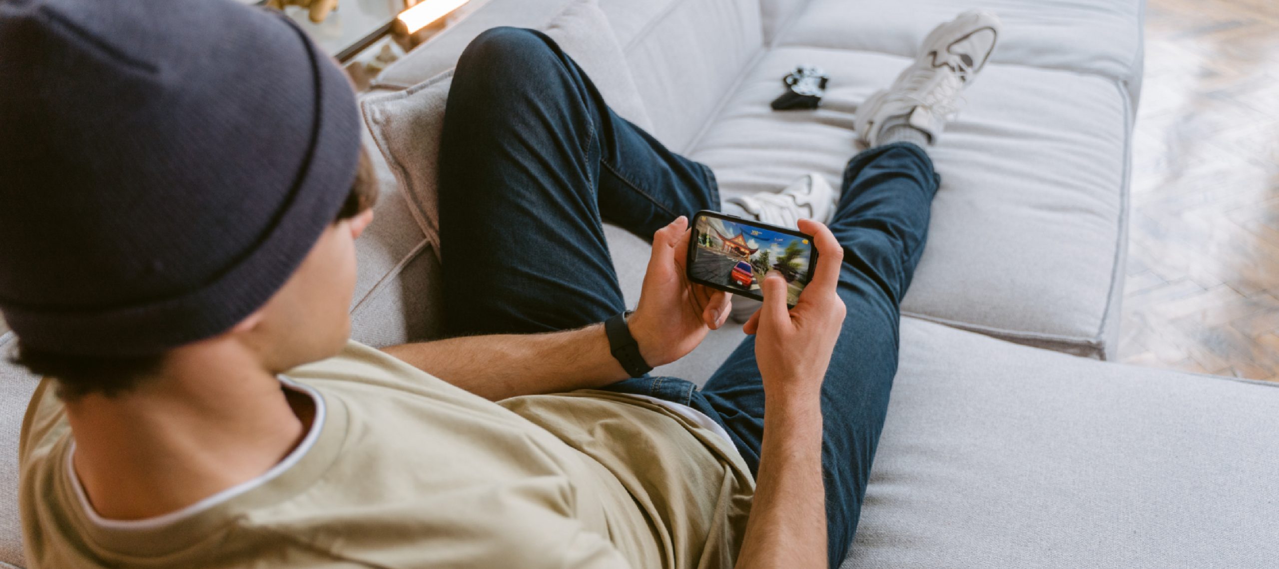 large image of man with a hat on inside playing a mobile game on his sofa