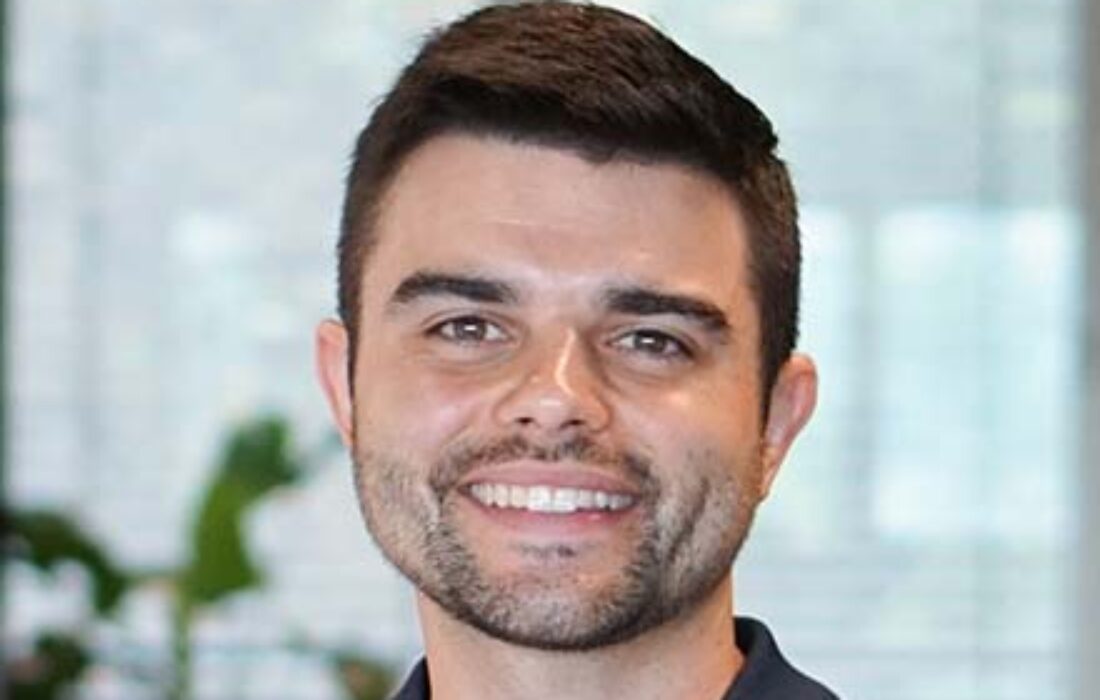 young dark-haired man with office behind him smiling at camera